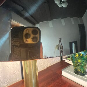 Puede incluir: Una l&aacute;mpara de mesa de color lat&oacute;n con una pantalla redonda y reflectante. La l&aacute;mpara tiene una base cil&iacute;ndrica y una mesa de madera marr&oacute;n oscuro. El reflejo muestra un techo negro y un tel&eacute;fono en una funda negra.