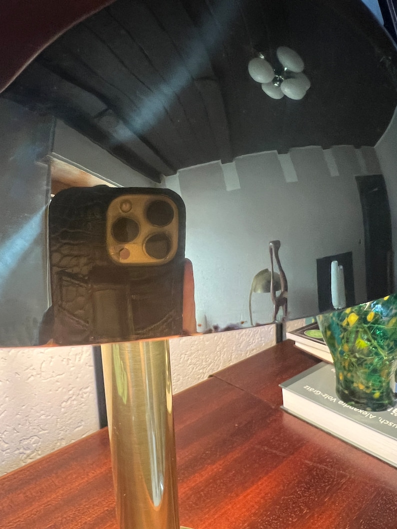 Puede incluir: Una l&aacute;mpara de mesa de color lat&oacute;n con una pantalla redonda y reflectante. La l&aacute;mpara tiene una base cil&iacute;ndrica y una mesa de madera marr&oacute;n oscuro. El reflejo muestra un techo negro y un tel&eacute;fono en una funda negra.