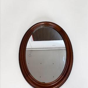 May include: Oval-shaped wooden framed mirror. The frame is a rich, dark brown color with a glossy finish. The mirror reflects the surrounding environment. The mirror is mounted on a white wall.