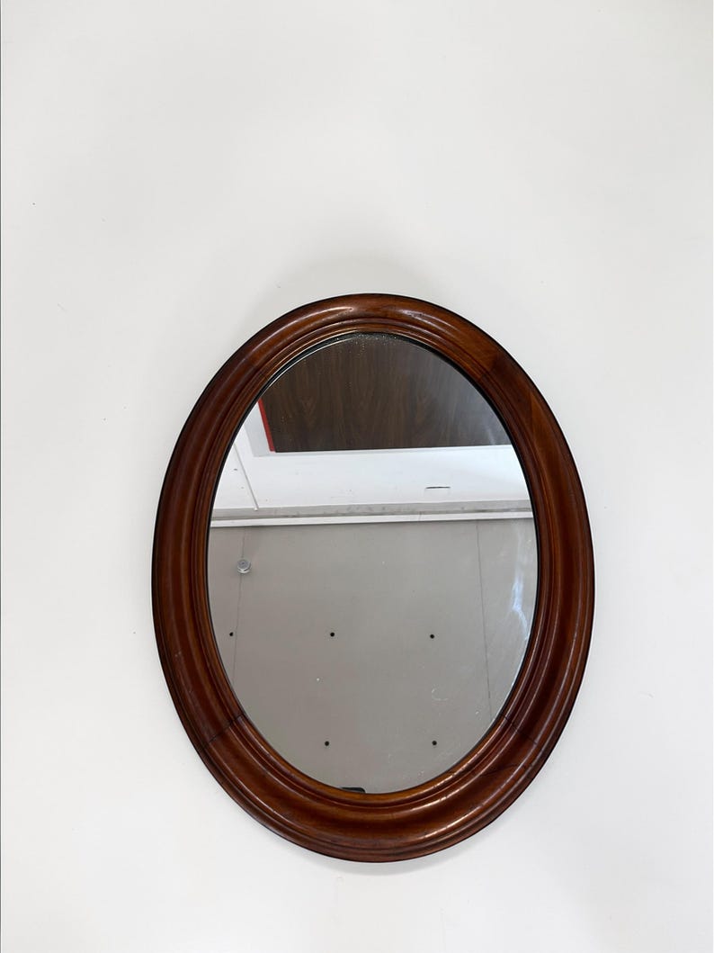 May include: Oval-shaped wooden framed mirror. The frame is a rich, dark brown color with a glossy finish. The mirror reflects the surrounding environment. The mirror is mounted on a white wall.