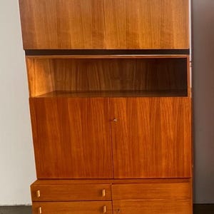 Puede incluir: Un mueble de madera de mediados de siglo con un acabado c&aacute;lido marr&oacute;n rojizo. El mueble presenta una secci&oacute;n superior con dos puertas, una estanter&iacute;a abierta, un armario central con puertas y una secci&oacute;n inferior con cajones. El dise&ntilde;o tiene una parte superior inclinada &uacute;nica.