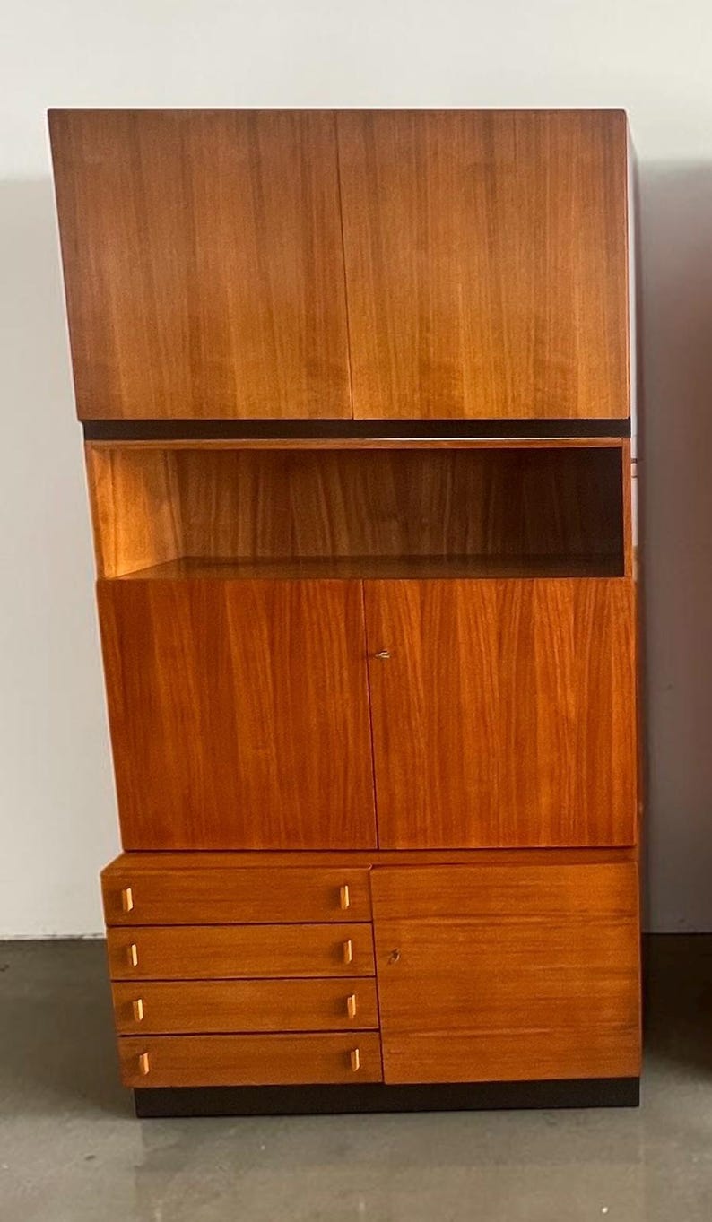 Puede incluir: Un mueble de madera de mediados de siglo con un acabado c&aacute;lido marr&oacute;n rojizo. El mueble presenta una secci&oacute;n superior con dos puertas, una estanter&iacute;a abierta, un armario central con puertas y una secci&oacute;n inferior con cajones. El dise&ntilde;o tiene una parte superior inclinada &uacute;nica.