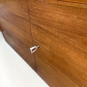 Puede incluir: Primer plano de un mueble de madera con un acabado de vetas naturales. Las puertas del mueble est&aacute;n cerradas, con una llave plateada insertada en la cerradura. La madera tiene un tono marr&oacute;n rojizo c&aacute;lido.