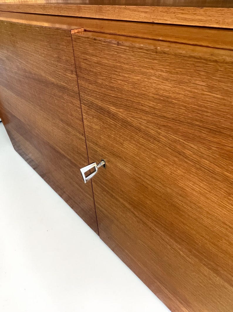 Puede incluir: Primer plano de un mueble de madera con un acabado de vetas naturales. Las puertas del mueble est&aacute;n cerradas, con una llave plateada insertada en la cerradura. La madera tiene un tono marr&oacute;n rojizo c&aacute;lido.