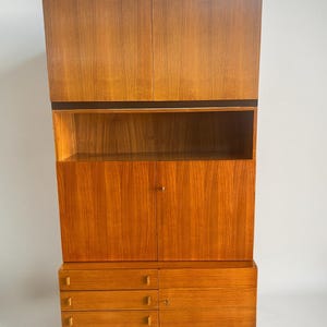 Puede incluir: Un mueble alto de madera de estilo moderno de mediados de siglo, con un acabado c&aacute;lido de color marr&oacute;n rojizo. El mueble presenta una combinaci&oacute;n de almacenamiento cerrado con puertas y cajones, y estanter&iacute;as abiertas. El dise&ntilde;o incluye una base y molduras de color marr&oacute;n oscuro, y una secci&oacute;n superior de dos puertas.