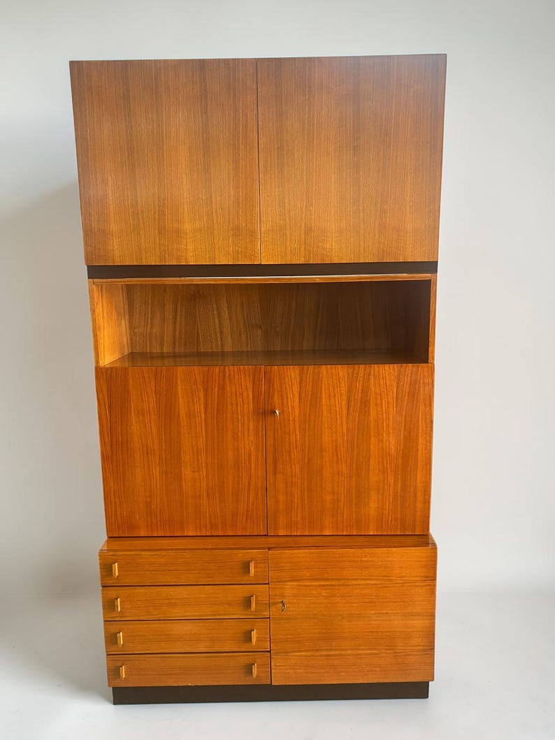 Puede incluir: Un mueble alto de madera de estilo moderno de mediados de siglo, con un acabado c&aacute;lido de color marr&oacute;n rojizo. El mueble presenta una combinaci&oacute;n de almacenamiento cerrado con puertas y cajones, y estanter&iacute;as abiertas. El dise&ntilde;o incluye una base y molduras de color marr&oacute;n oscuro, y una secci&oacute;n superior de dos puertas.