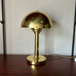 Puede incluir: Una l&aacute;mpara de escritorio de lat&oacute;n con una pantalla en forma de c&uacute;pula y una base cil&iacute;ndrica. La l&aacute;mpara tiene un acabado pulido y un dise&ntilde;o simple y elegante. La l&aacute;mpara mide aproximadamente 30 cm de altura.