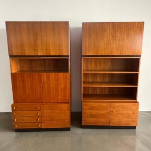 Puede incluir: Dos armarios de madera a juego con un acabado marr&oacute;n rojizo c&aacute;lido. Uno tiene cajones y puertas, el otro estantes abiertos. Ambos tienen una banda oscura en la parte superior y se asientan sobre una base oscura. Los armarios son altos y rectangulares.