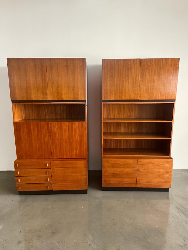Puede incluir: Dos armarios de madera a juego con un acabado marr&oacute;n rojizo c&aacute;lido. Uno tiene cajones y puertas, el otro estantes abiertos. Ambos tienen una banda oscura en la parte superior y se asientan sobre una base oscura. Los armarios son altos y rectangulares.