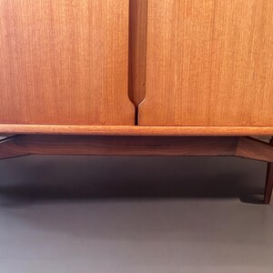 Puede incluir: Un armario de madera vintage con dos puertas y un estante inferior. El armario tiene un acabado de madera clara y se apoya en cuatro patas anguladas.