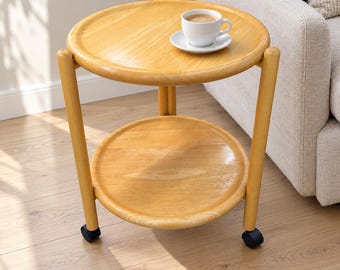 Puede incluir: Una mesa auxiliar redonda de madera clara con dos niveles y cuatro ruedas negras. El nivel superior sostiene una taza de café blanca con su plato. La mesa presenta un acabado liso y pulido con un diseño sencillo y funcional.