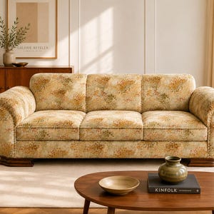 May include: A three-cushion sofa upholstered in a light beige fabric featuring a dense, multi-coloured floral pattern in shades of orange, yellow, and green. The sofa has rounded arms and is supported by dark brown wooden feet at the base.