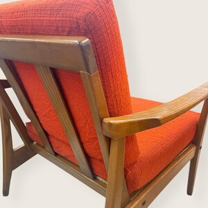May include: Mid-century modern armchair with a wooden frame and vibrant orange upholstery. The chair features a slatted back and armrests, showcasing a blend of wood and fabric textures. A stylish piece of furniture.