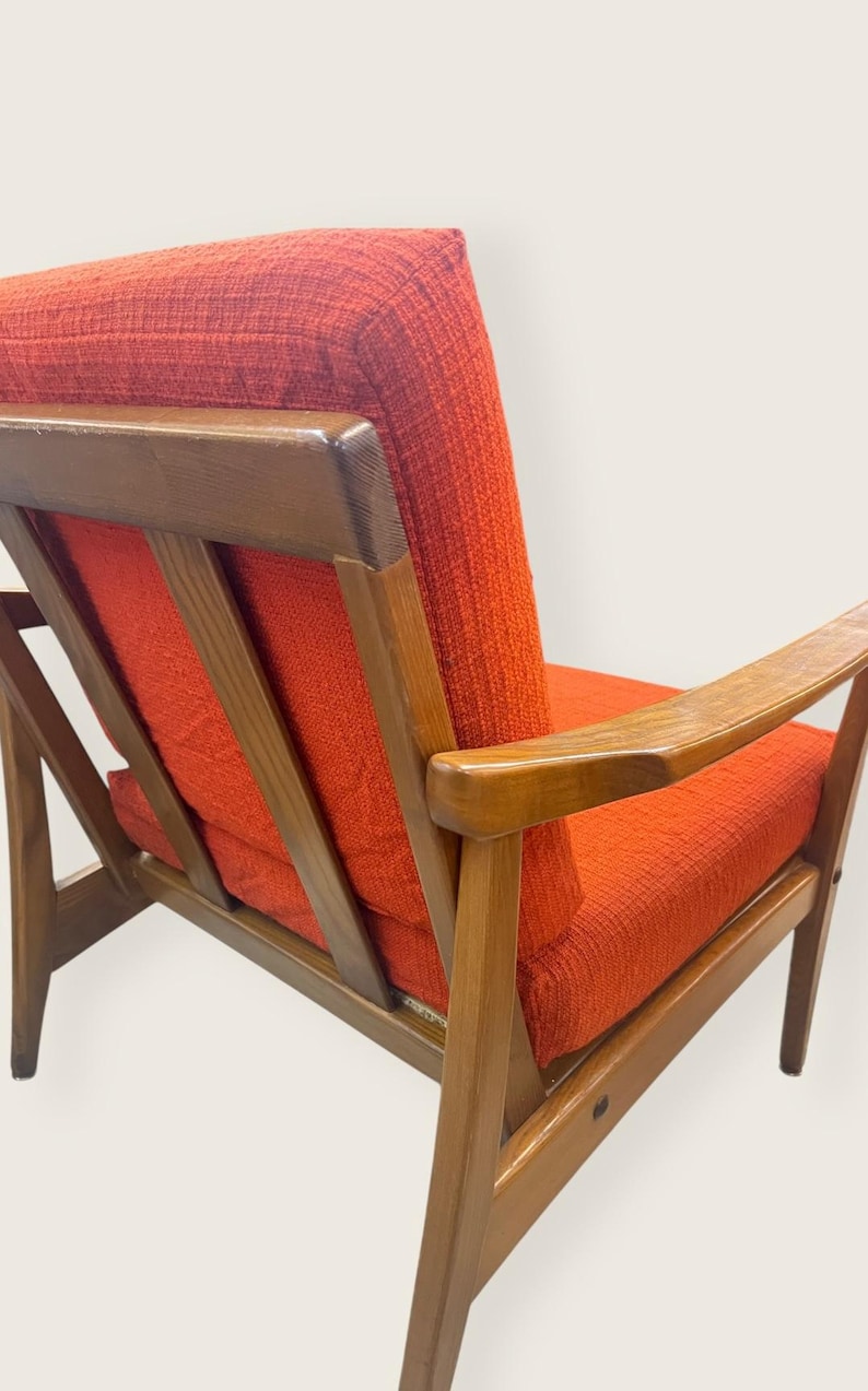 May include: Mid-century modern armchair with a wooden frame and vibrant orange upholstery. The chair features a slatted back and armrests, showcasing a blend of wood and fabric textures. A stylish piece of furniture.