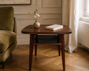 May include: A dark brown wooden side table with a square top featuring rounded corners and a lower shelf. A small beige ceramic vase containing white flowers and a closed rectangular book with a white cover are placed on the table surface.