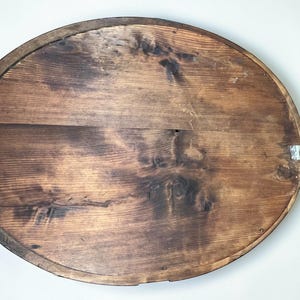 May include: Oval wooden wall art with a dark brown finish. The back of the piece shows the wood grain and a metal hanger. The wood has a distressed look with knots and variations in color.