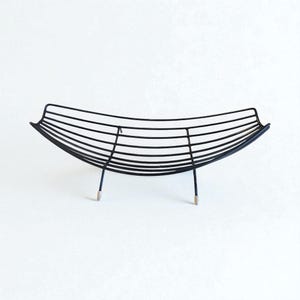 Op de afbeelding: Een zwarte metalen draad fruitmand met een gebogen, bootachtige vorm. De mand heeft een reeks horizontale draden die worden ondersteund door vier dunne poten met zilverkleurige uiteinden. Minimalistisch en modern ontwerp.