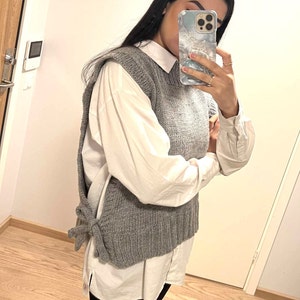 May include: A gray knitted vest with a tie at the back, worn over a white button-down shirt.