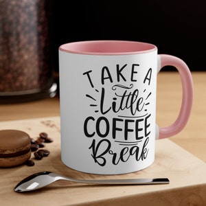 Puede incluir: Taza de café blanca con interior y asa rosas. La taza presenta el texto "TAKE A Little COFFEE Break" en escritura negra. Una cuchara, granos de café y un macaron están sobre una superficie de madera.