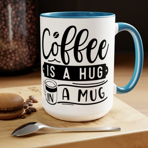 Könnte beinhalten: Eine weiße Kaffeetasse mit blauem Rand und Henkel. Die Tasse hat einen schwarzen Schriftzug mit der Aufschrift "Coffee is a hug in a mug".