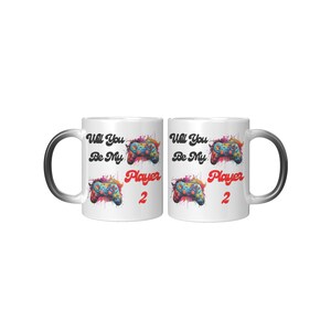 May include: Two white ceramic mugs with black handles. Both mugs feature a colorful video game controller graphic with the text "Will You Be My Player 2" in black and red.