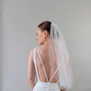 Peut inclure: Un voile de mariée blanc à pois. Le voile est fixé aux cheveux et retombe sur l'épaule. La mariée porte une robe blanche avec un dos nu.