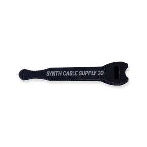 May include: Black cable tie with the text "SYNTH CABLE SUPPLY CO" in white. The tie has a rounded end and a notched end with a slot. The cable tie is designed to organize and secure cables.