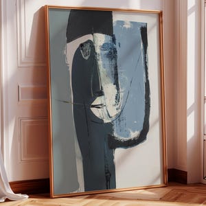 May include: Abstract art print featuring a stylized face in shades of blue, gray, and white. The artwork is framed in a gold-colored frame and leans against a white wall. The artwork's style is modern and minimalist.