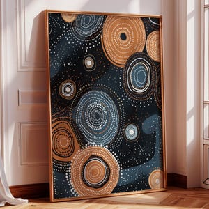 May include: Abstract art print with a pattern of concentric circles in navy, tan, and white against a dark background. The artwork is framed in a gold-coloured frame and leans against a white wall.