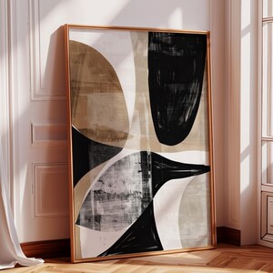 Puede incluir: Impresión artística abstracta en un marco dorado. La obra presenta una composición de formas negras, blancas y beige. Las formas incluyen formas curvas y geométricas, creando una estética moderna y minimalista. El arte enmarcado está apoyado contra una pared blanca.