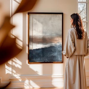 May include: An abstract landscape painting in a dark wooden frame. The artwork features a blend of colors, including shades of blue, brown, and white. The painting hangs on a cream-colored wall. A person is standing in front of the painting.