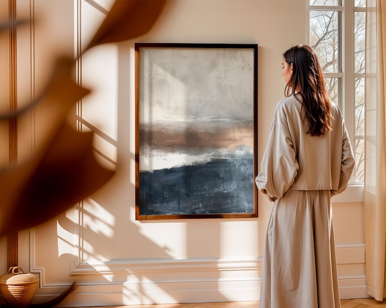 May include: An abstract landscape painting in a dark wooden frame. The artwork features a blend of colors, including shades of blue, brown, and white. The painting hangs on a cream-colored wall. A person is standing in front of the painting.