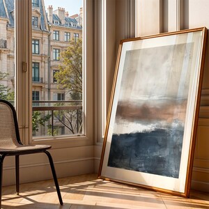 May include: An abstract landscape painting in a gold frame, leaning against a wall. The artwork features a blend of brown, beige, and blue hues. A black chair with a woven back sits near a window overlooking a city.
