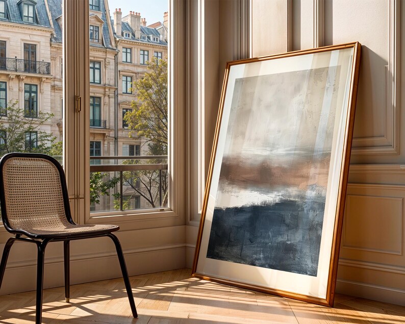 May include: An abstract landscape painting in a gold frame, leaning against a wall. The artwork features a blend of brown, beige, and blue hues. A black chair with a woven back sits near a window overlooking a city.