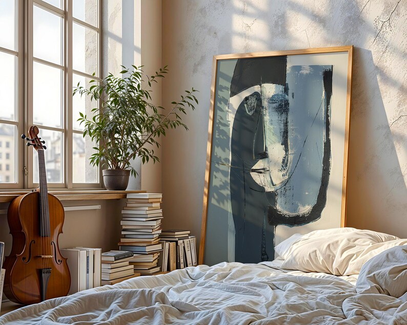 May include: A cello stands near a window with a stack of books and a potted plant. A large framed abstract art piece leans against a bed with a white comforter. The artwork features a face in shades of blue and gray.