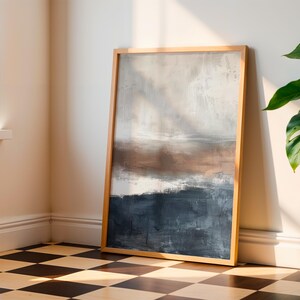 May include: Abstract art print in a natural wood frame. The artwork features a landscape-style composition with a color palette of white, brown, and navy blue. The framed art is leaning against a wall in a room with a checkered floor.