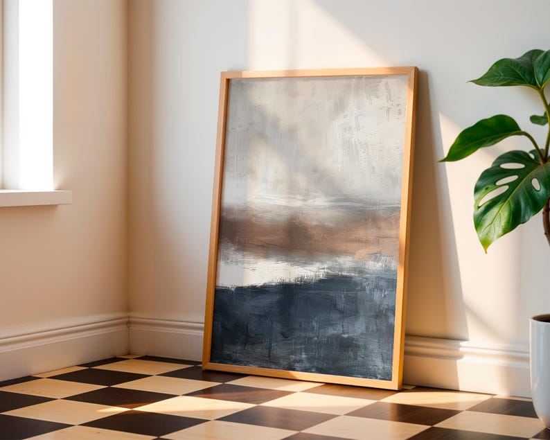 May include: Abstract art print in a natural wood frame. The artwork features a landscape-style composition with a color palette of white, brown, and navy blue. The framed art is leaning against a wall in a room with a checkered floor.