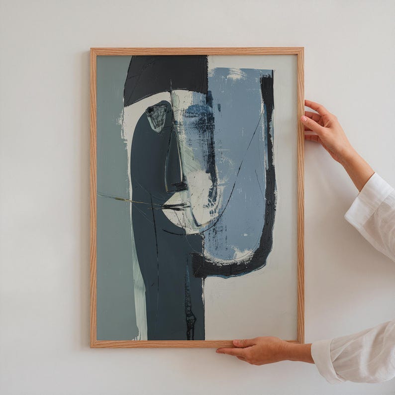 May include: Abstract face art print in a natural wood frame. The artwork features a split design with shades of blue, gray, and black. The face is depicted with minimalist lines and shapes, creating a modern aesthetic.