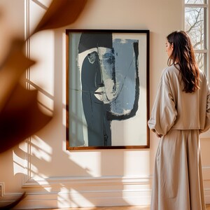 May include: Abstract art print in a dark wooden frame. The artwork features a face with blue, gray, and white brushstrokes. The painting hangs on a cream-colored wall. A woman in a beige outfit stands near the artwork.
