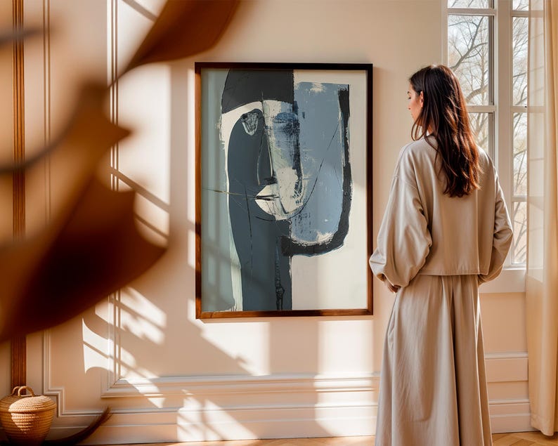 May include: Abstract art print in a dark wooden frame. The artwork features a face with blue, gray, and white brushstrokes. The painting hangs on a cream-colored wall. A woman in a beige outfit stands near the artwork.