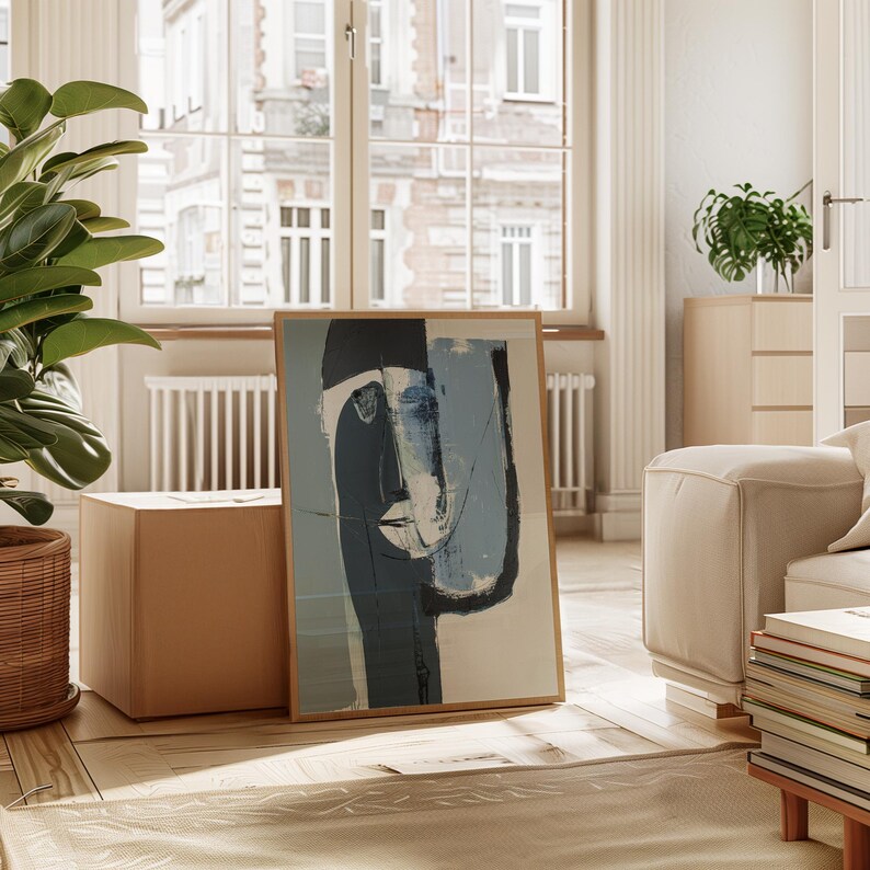 May include: Abstract art print featuring a stylized face in shades of blue, black, and white, framed in natural wood. The artwork leans against a neutral-toned wall, adding a touch of modern art to the interior decor.