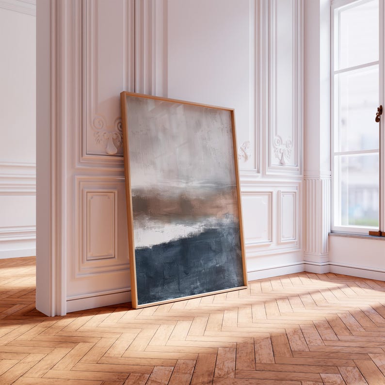 May include: A framed abstract painting with a wooden frame. The artwork features a landscape-style composition with a color palette of gray, brown, white, and navy blue. The painting is leaning against a white wall in a room with a wooden floor.