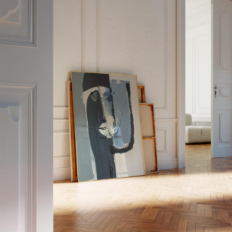 May include: Abstract art print featuring a stylized face in shades of gray, blue, and black, framed with a gold border. The artwork leans against a white wall, creating a minimalist aesthetic. The room has a wooden floor.