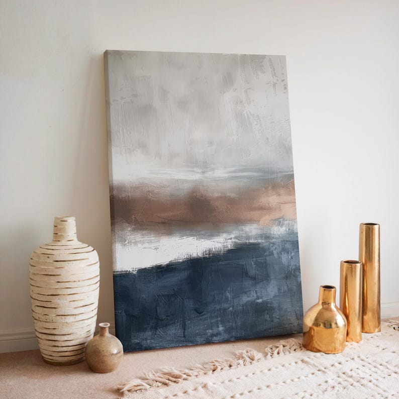 May include: Abstract landscape painting with a color palette of white, gray, brown, and navy blue. The artwork is leaning against a white wall, with decorative vases in the foreground. The vases include a large, textured white vase, a small brown vase, and three gold-colored vases.
