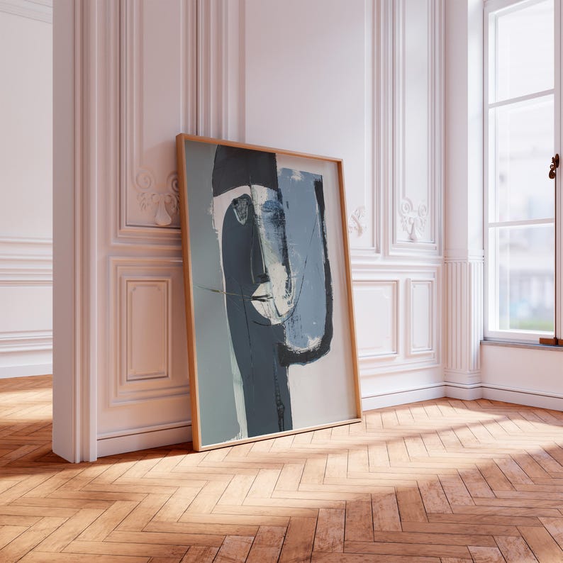 May include: Abstract art print featuring a stylized face in shades of blue, gray, and black. The artwork is framed in a light wood frame and leans against a white wall with decorative molding. The floor is made of wooden planks.