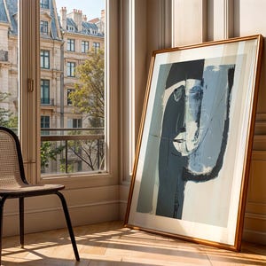 May include: An abstract art print in a wooden frame, leaning against a wall. The artwork features a face-like composition in shades of blue, gray, and white. A black chair with a woven seat sits near a window overlooking a city.
