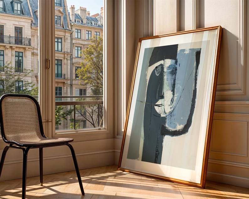 May include: An abstract art print in a wooden frame, leaning against a wall. The artwork features a face-like composition in shades of blue, gray, and white. A black chair with a woven seat sits near a window overlooking a city.