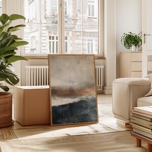 May include: Abstract landscape art print in a wooden frame. The artwork features a color palette of beige, brown, and navy blue. The painting is leaning against a wall in a well-lit room with a window in the background. The style is modern and minimalist.