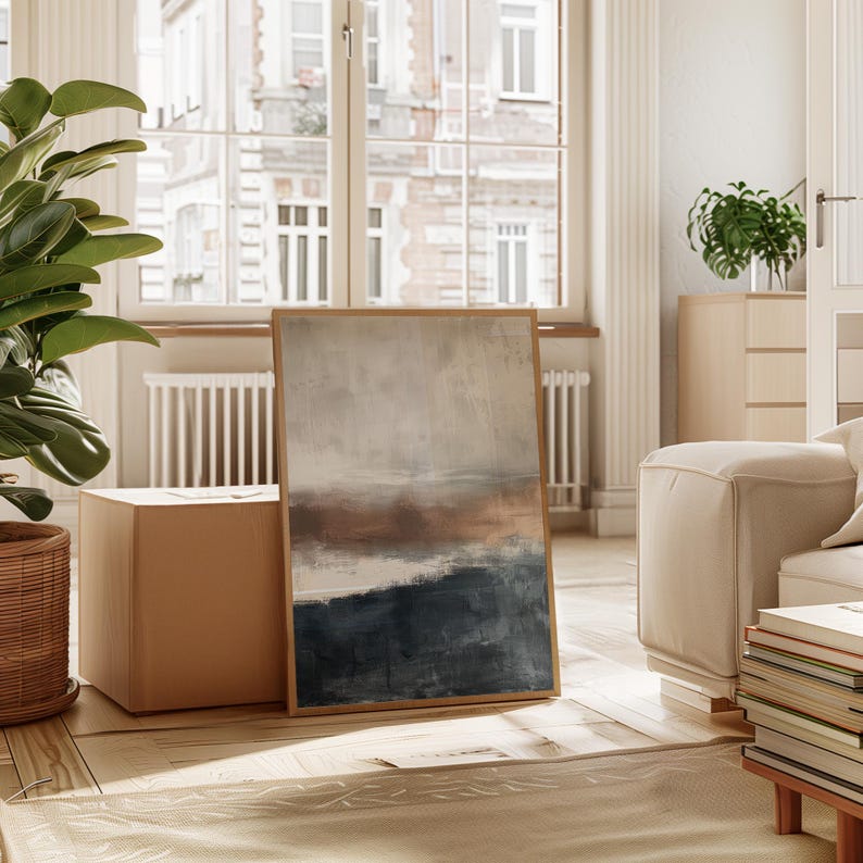 May include: Abstract landscape art print in a wooden frame. The artwork features a color palette of beige, brown, and navy blue. The painting is leaning against a wall in a well-lit room with a window in the background. The style is modern and minimalist.