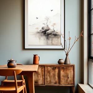May include: A framed watercolor painting of a small island with trees and birds in a misty landscape. The painting is hanging on a light green wall above a wooden cabinet with two chairs and a table in front of it.